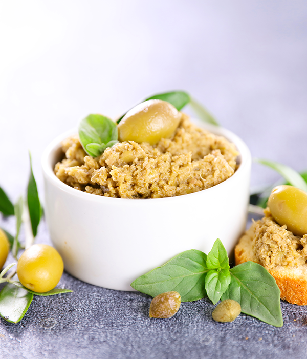 A Vertical Image Featuring a Small White Bowl Filled with Tapenade. A Full Green Olive Sits on Top of and Around the Bowl.