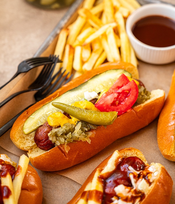 Close up of Chicago Dog (topped with yellow mustard, sweet relish, white onion, tomato, a dill pickle, a Sport pepper, and a dash of celery sauce) with fries and BBQ sauce in the background