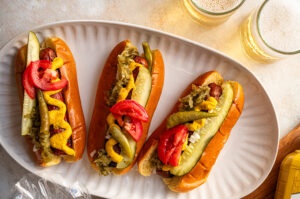 Three Chicago Hotdogs (topped with yellow mustard, sweet relish, white onion, tomato, a dill pickle, a Sport pepper, and a dash of celery sauce) on a paper plate