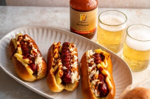 Three Cleveland Hotdogs (topped with fries, cole slaw, and BBQ sauce) on a paper plate with two glasses of beer in the background