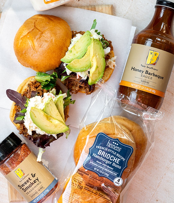 Hatch Chili Pulled Pork Sandwich on a wooden board covered with parchment paper, and surrounded by Two Brother's BBQ Sauce and Sweet and Smokey Seasoning, and Heinen's Brioche Hamburger Buns
