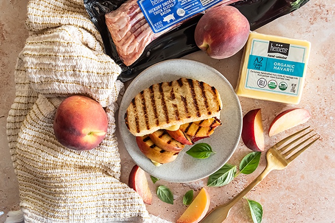 Peach grilled cheese with bacon on a plate surrounded by packaged bacon, fresh peaches, and Heinen's Organic Havarti cheese