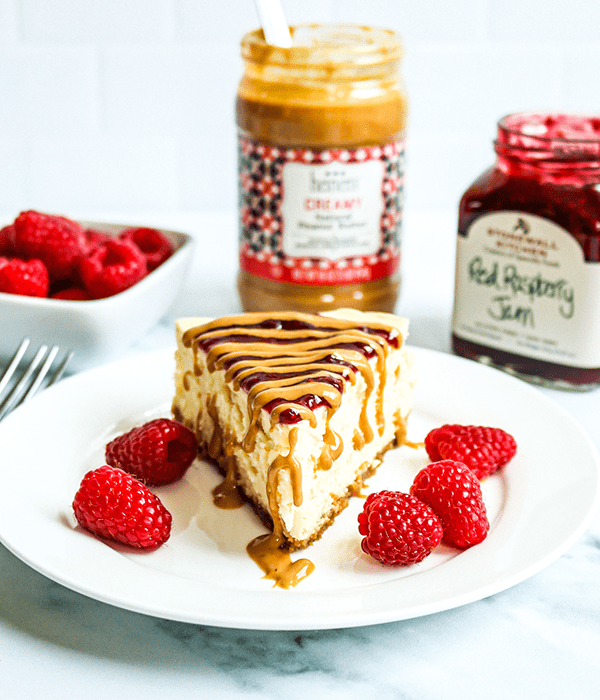 A Slice of Heinen's Original Cheesecake on a Plate with a Peanut Butter and Jelly Topping and Fresh Raspberries. A Jar of Peanut Butter, a Jar of Jam and a bowl of raspberries sit in the background.