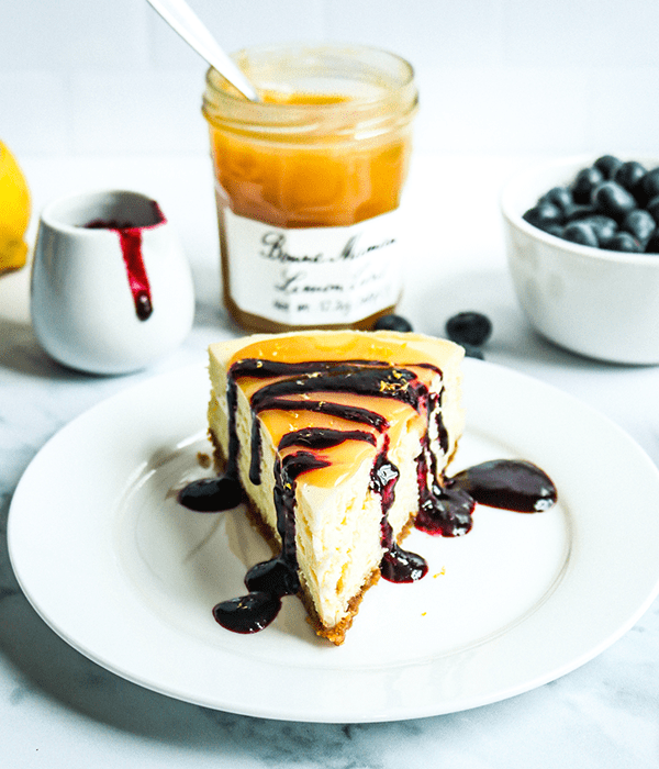 A Slice of Heinen's Original Cheesecake on a Plate with a Lemon Blueberry Topping. A Jar of Lemon Curd, a Jar of Blueberry Sauce and a bowl of blueberries sit in the background.
