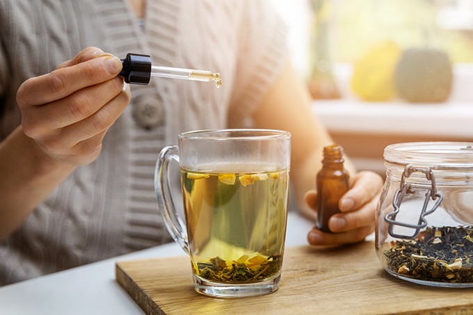 Woman adding drop of CBD oil in cup of tea with pipette