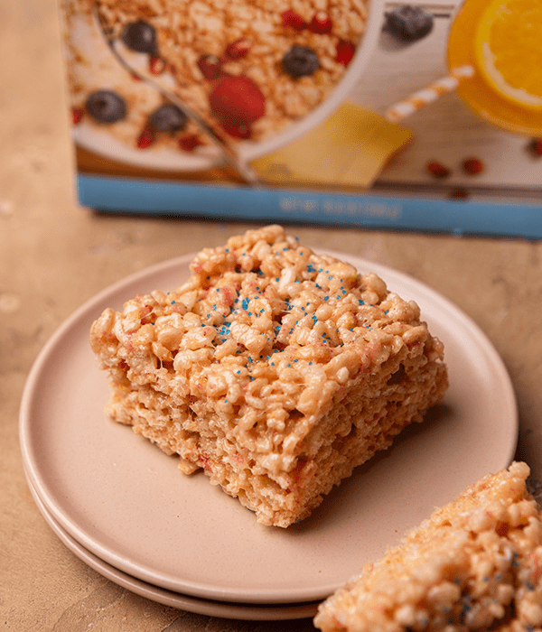 A Strawberry Crunch Rice Crispie Treat on a Plate in Front of a Box of Heinen's Rice Crispies Cereal