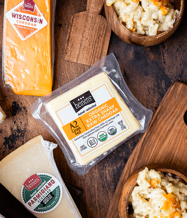 Three Packages of Heinen's Hand-Selected Cheeses on a Dark Wood Surface Beside Small Bowls of Macaroni and Cheese