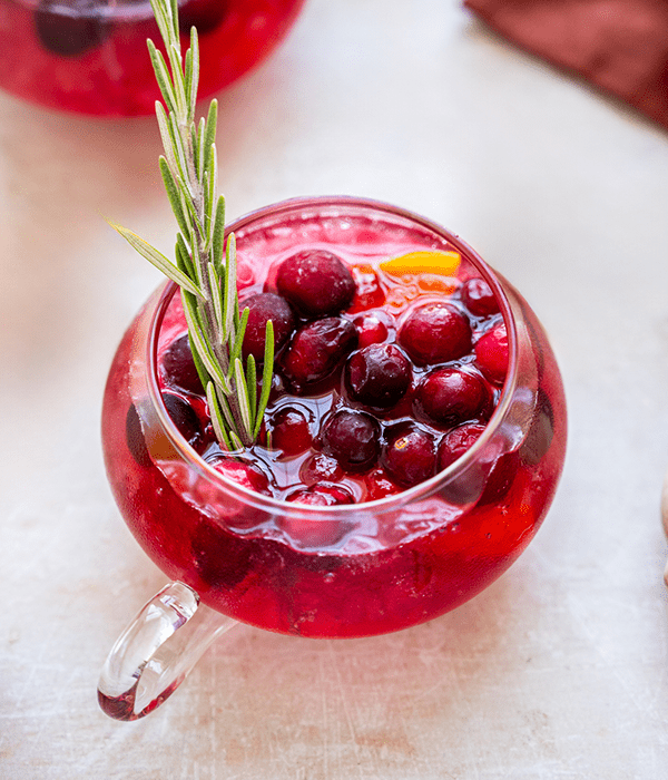 A Circular Glass Mug Filled with a Red Old Fashioned Holiday Punch with Garnishes of Cranberries, Rosemary Sprigs and Orange Slices