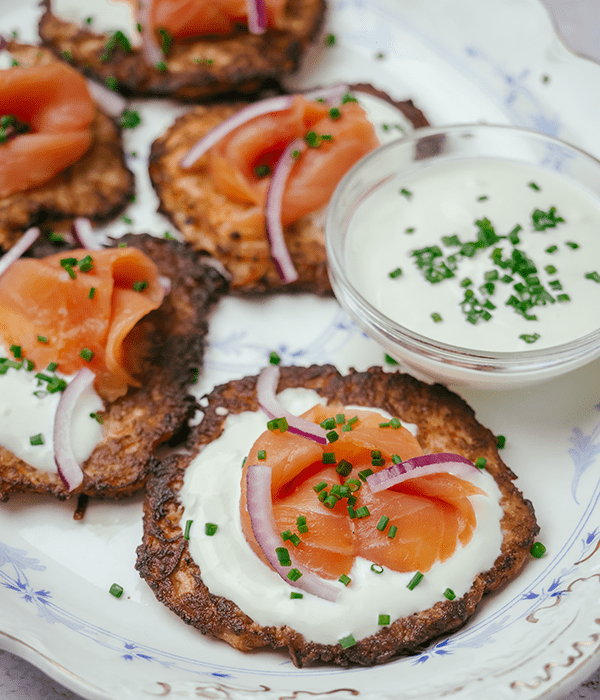 A Close Up Image of a Potato Pancake Topped with Creme Fraiche, Smoked Salmon, Red Onion, and Chives